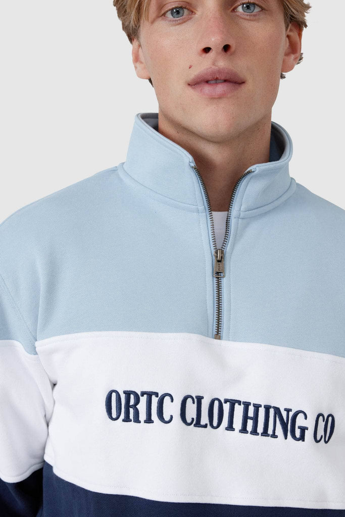 Male model wearing quarter zip jumper that has a light blue top panel, white middle panel with navy embroidered ortc Clothing Co and navy base panel